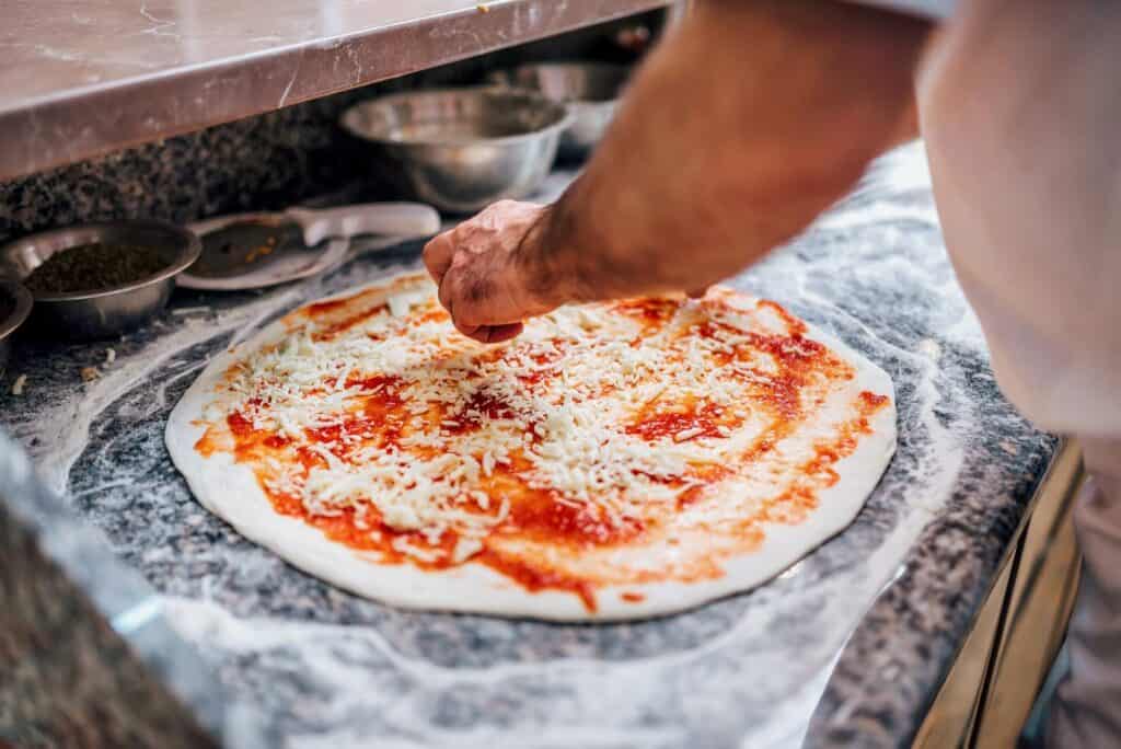 A chef adding cheese to an uncooked pizza with tomato