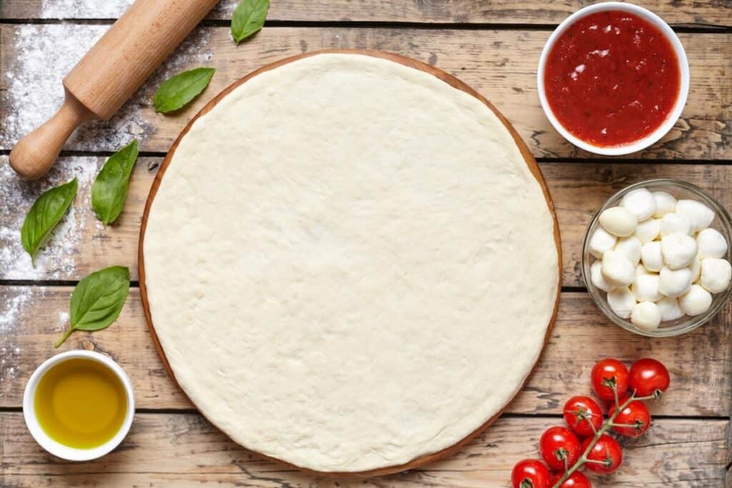 Pizza dough on the pizza stone, oil, cherry tomatoes, tomato sauce, and cheese next to it